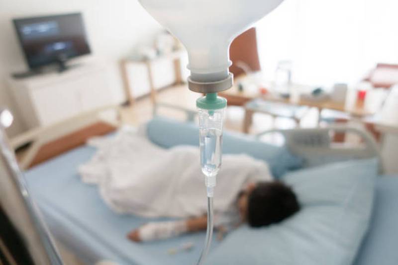 Foto de primer plano de un gotero de suero y de fondo la silueta de un niño en una cama de hospital.